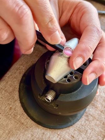 Close-up of hands using tweezers to set a pink cabochon gemstone into a silver bezel on a jeweler’s bench vise, handmade jewelry making scene.