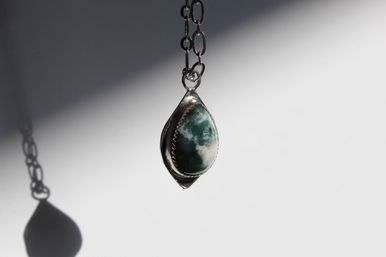 Close-up of an earthy green teardrop moss agate pendant set in silver, hanging from a chain and casting a crisp shadow on a white studio background.
