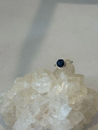 Close-up of a silver gemstone ring with a dark blue round stone perched on a sparkling clear quartz crystal cluster against a soft neutral background.