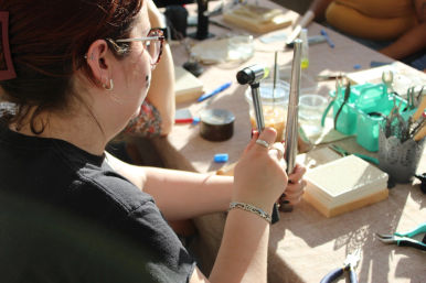 Person at a sunlit craft table shaping metal on a ring mandrel with a small hammer, surrounded by pliers, trays and jewelry-making supplies.
