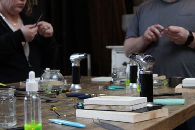 Close-up of an artisan workbench in a jewelry-making workshop with handheld butane torches, pliers, files, ceramic blocks and small glass jars, two crafters working in the background.