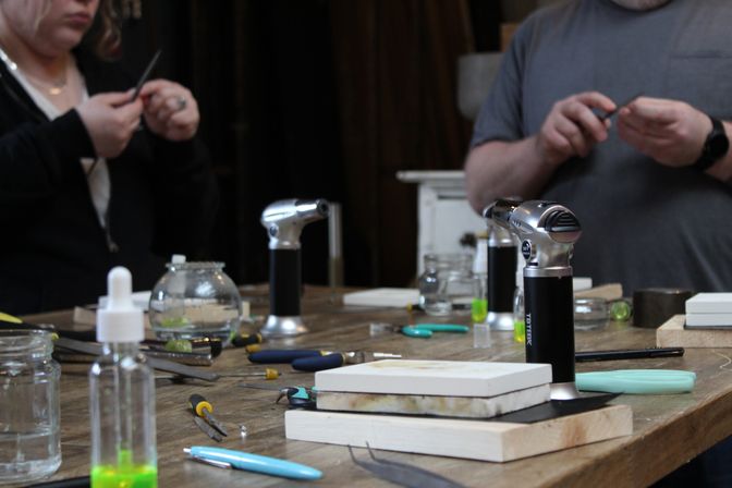 Close-up of an artisan workbench in a jewelry-making workshop with handheld butane torches, pliers, files, ceramic blocks and small glass jars, two crafters working in the background.