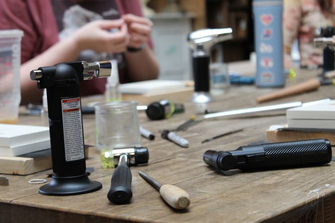 Busy wooden workbench in a craft workshop with a tabletop butane torch, handheld torch, mallet, awl, markers, small jars and blurred hands working in the background.