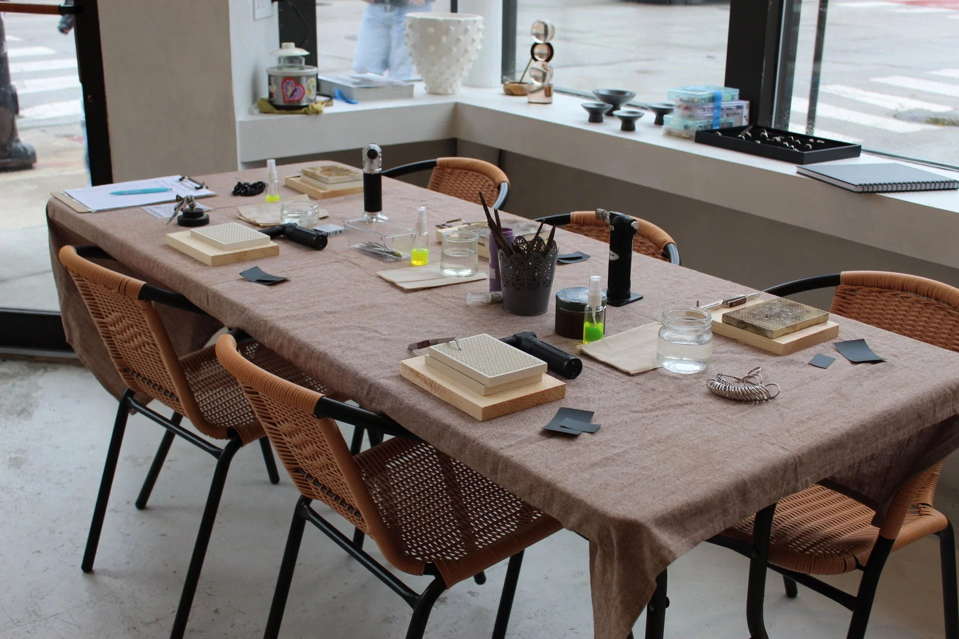 Communal jewelry-making workshop table set with wooden blocks, mesh pads, small torches, jars of water, tools and woven chairs beside a street-facing studio window