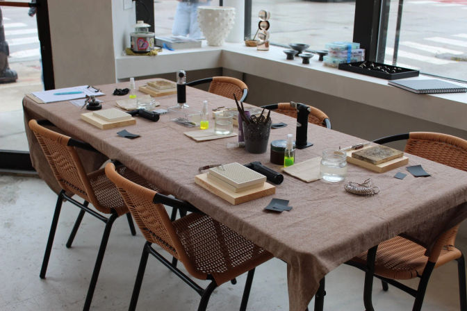 Communal jewelry-making workshop table set with wooden blocks, mesh pads, small torches, jars of water, tools and woven chairs beside a street-facing studio window