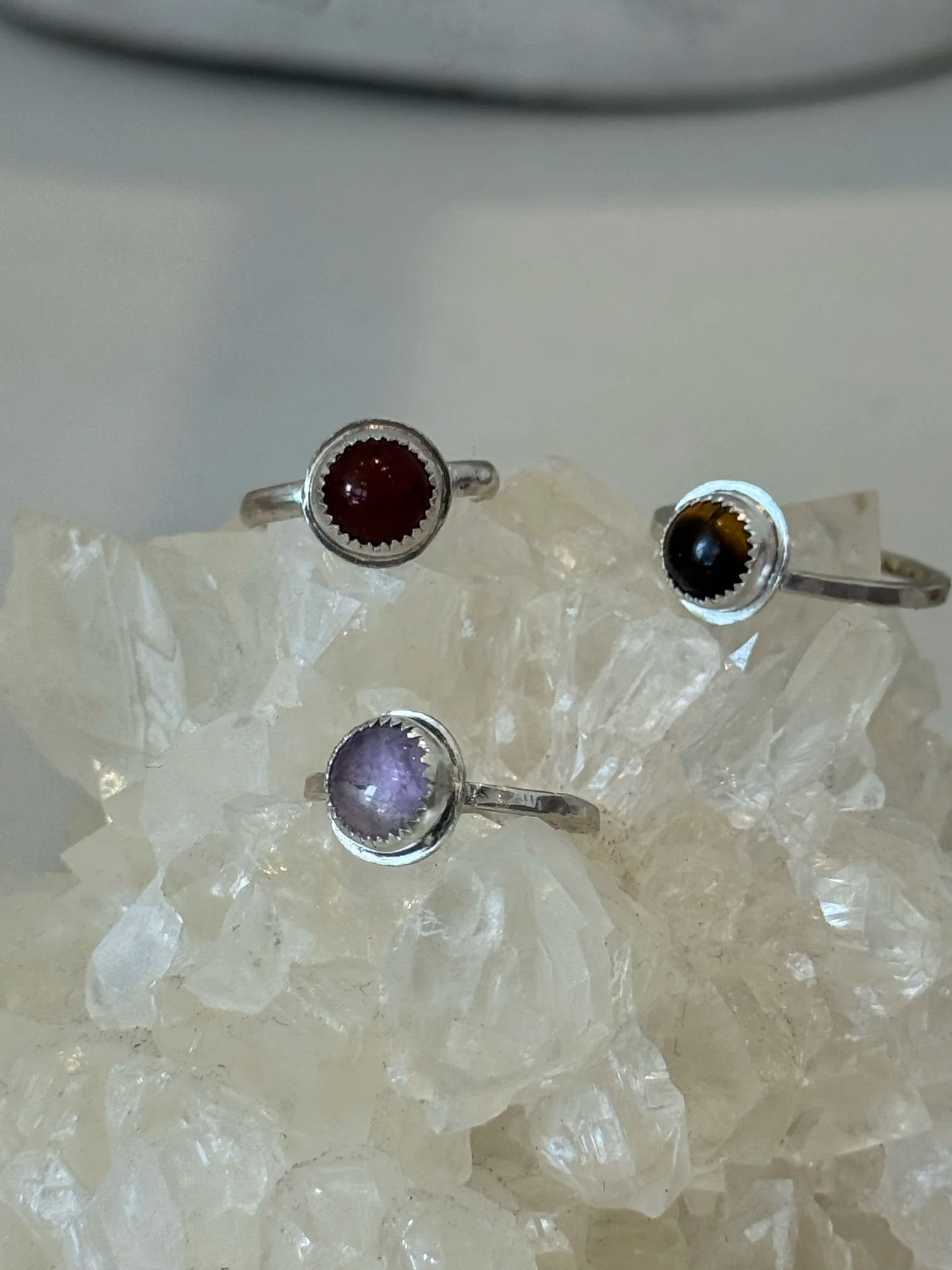 Three dainty sterling silver rings with purple, red, and brown cabochon stones displayed on a white quartz crystal cluster.