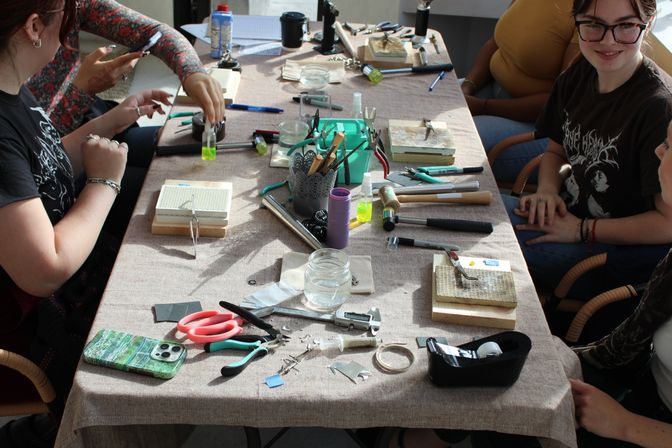 Sunlit craft workshop table with jewelry-making tools—pliers, hammers, mandrels, soldering blocks, small vials, tape and a phone—with students' hands working together around it