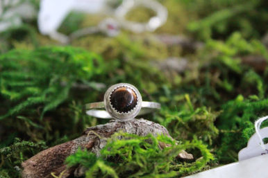 Close-up of a silver ring with a round brown tiger's eye gemstone resting on moss-covered bark — nature-inspired jewelry shot.