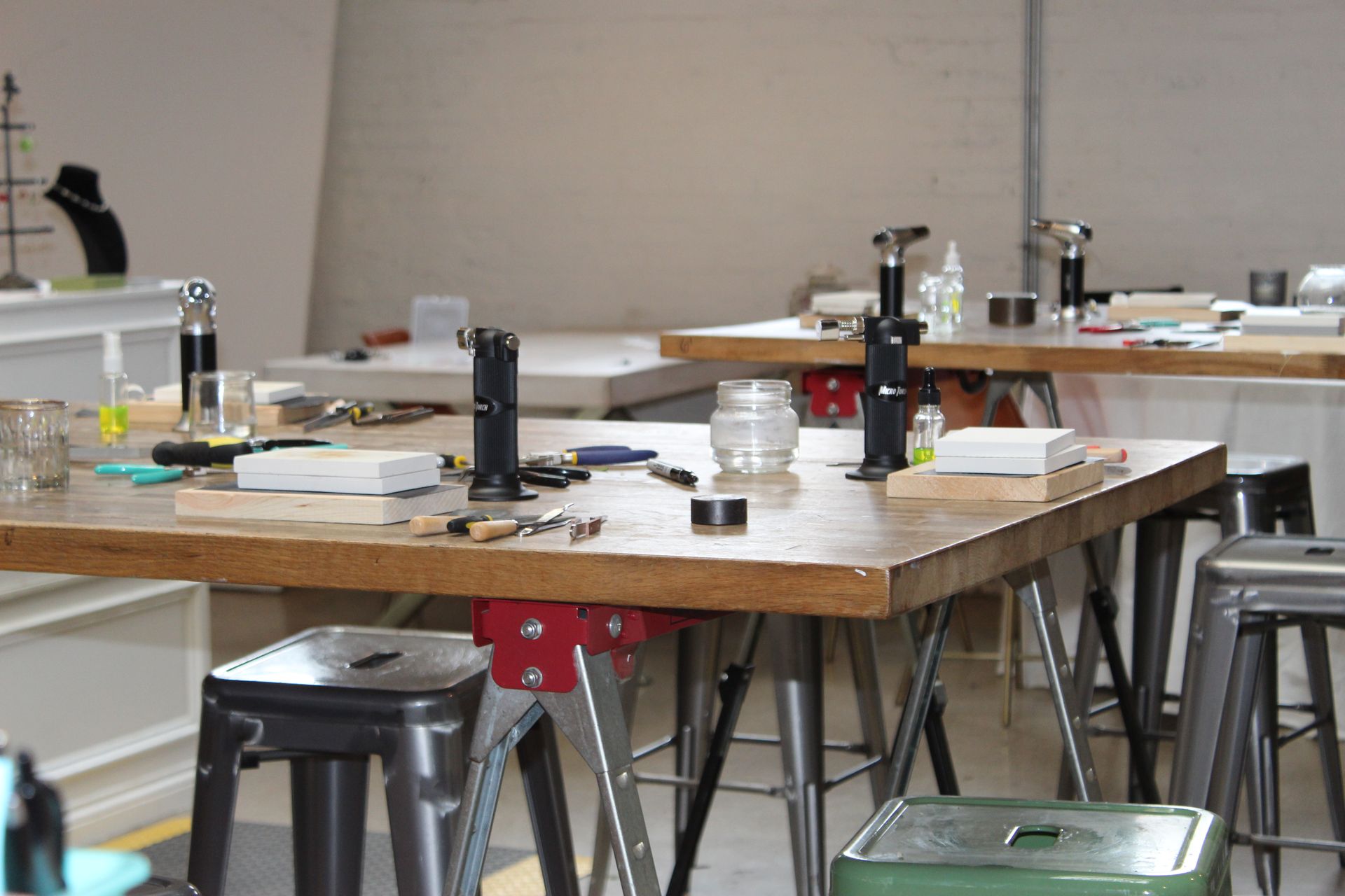Jewelry-making workshop workbench with tabletop torches, pliers, hand tools, wooden blocks, glass jars and metal stools in a bright studio