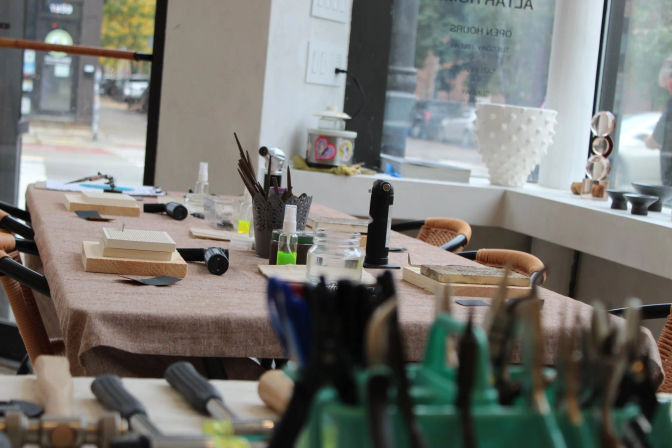 Light-filled craft workshop table by a storefront window, set with hand tools and pliers, wooden blocks, jars, and small torches for jewelry or metalworking projects.