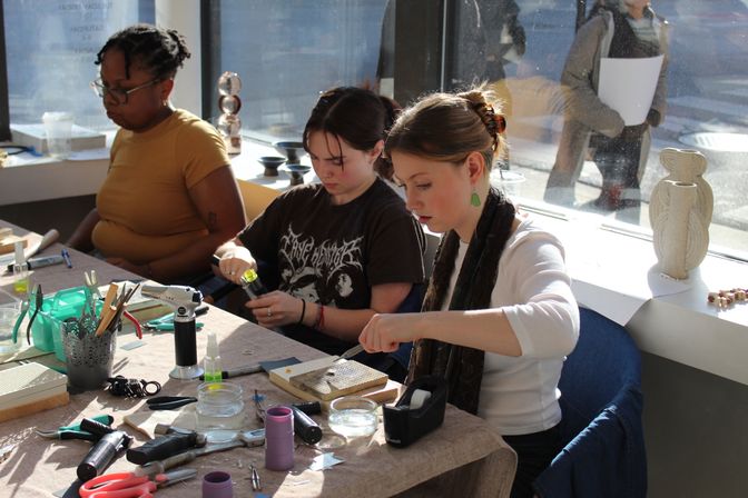 Sunlit craft workshop with three participants seated at a table by a large window, focused on jewelry making and metalwork using pliers, a handheld torch, files, jars, and other small tools.