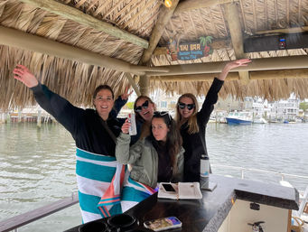 Four women smiling and waving under a thatched tiki bar on a boat at a marina, two wrapped in striped beach towels and holding drinks with docks and waterfront houses visible behind them.