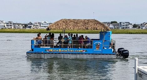 Blue party pontoon with a thatched tiki roof and outboard motor carrying a group of passengers on calm coastal water, with marsh grass and waterfront houses in the background.
