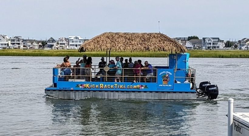 Blue party pontoon with a thatched tiki roof and outboard motor carrying a group of passengers on calm coastal water, with marsh grass and waterfront houses in the background.