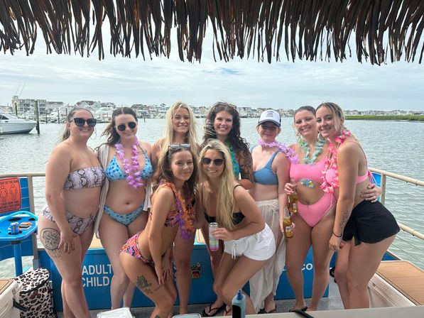 Nine friends in colorful swimsuits and leis smiling and posing on a tiki-roofed boat deck with drinks, docked at a coastal marina with boats and waterfront homes in the background.