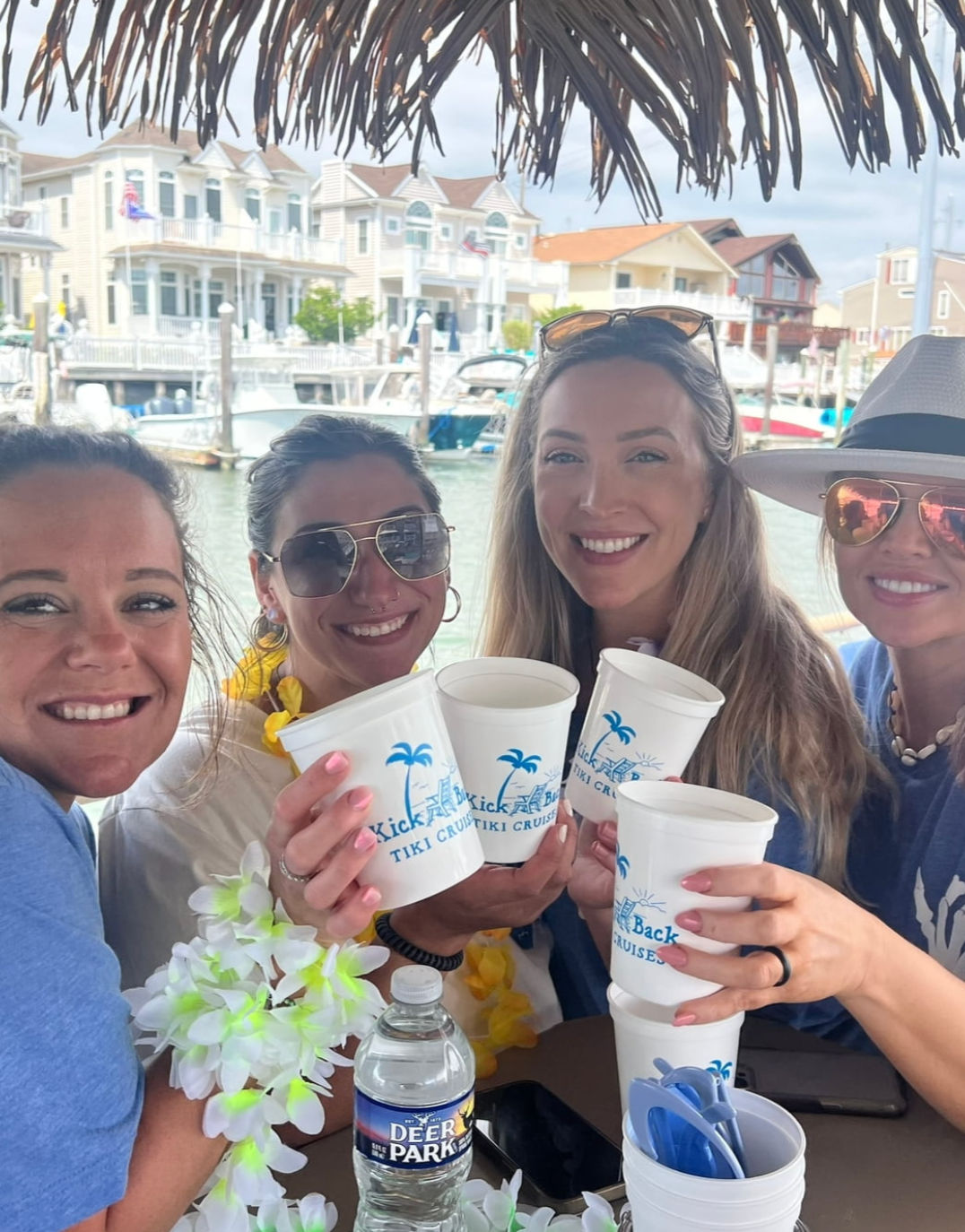 Four smiling friends clinking white tiki-style cups under a thatched umbrella at a marina, leis and sunglasses adding tropical flair, with boats and waterfront vacation homes in the background — summer boat cruise vibe.