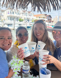 Four smiling friends clinking white tiki-style cups under a thatched umbrella at a marina, leis and sunglasses adding tropical flair, with boats and waterfront vacation homes in the background — summer boat cruise vibe.