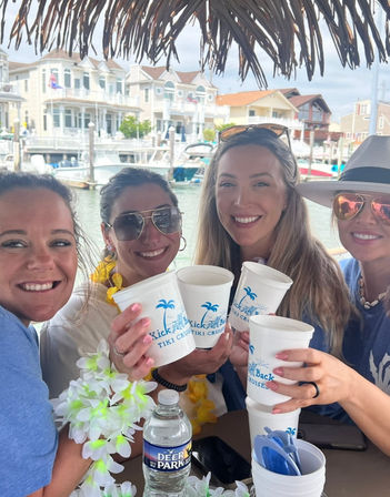 Four smiling friends clinking white tiki-style cups under a thatched umbrella at a marina, leis and sunglasses adding tropical flair, with boats and waterfront vacation homes in the background — summer boat cruise vibe.