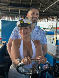 Smiling couple steering a boat at a sunny marina — woman wearing a white “bride” captain’s hat and halter top at the wheel, man in a blue floral shirt behind her, docked boats and waterfront in the background.