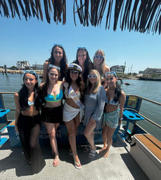 Eight friends in swimsuits and blue sunglasses pose on a boat in a sunny coastal harbor, celebrating a bachelorette with the bride-to-be wearing a captain’s hat and sash.