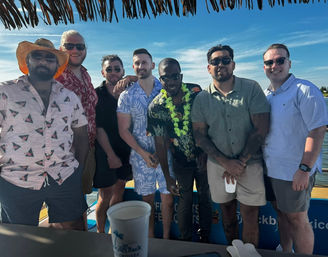 Seven friends in tropical shirts and sunglasses posing on a sunny boat cruise with a thatched-roof bar, a green lei, and blue coastal waters in the background — tropical vacation vibe.