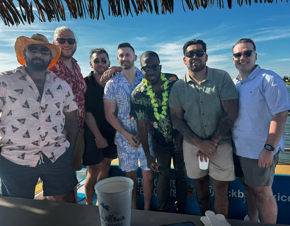 Seven friends in tropical shirts and sunglasses posing on a sunny boat cruise with a thatched-roof bar, a green lei, and blue coastal waters in the background — tropical vacation vibe.