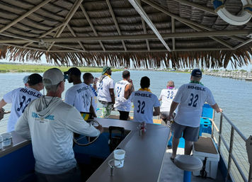 Group of people in matching 'BIMBLE 32' shirts on a tiki-roofed pontoon boat cruising a coastal marina and marshy inlet.