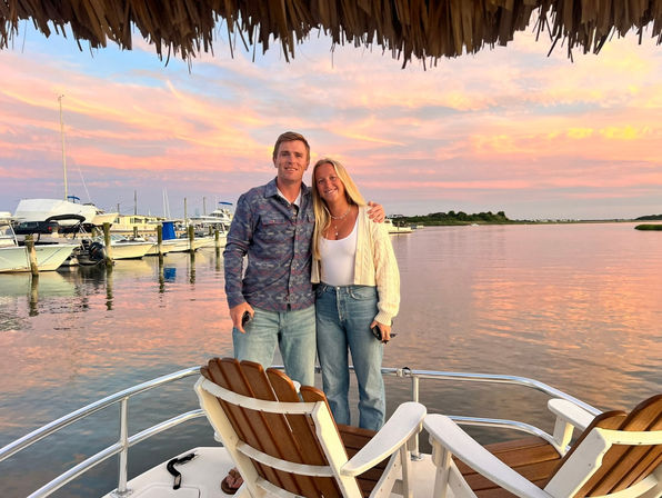 Couple standing on a boat deck beneath a thatched roof at a coastal marina during a pastel pink-orange sunset, with yachts docked nearby and calm water reflecting the sky.