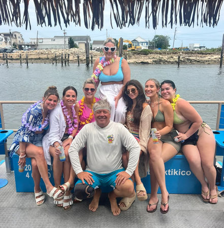 Smiling group of eight adults on a tiki-style party boat in a coastal harbor, women in swimsuits and colorful leis with a man in shorts kneeling in front, rocky shoreline, docks and seaside houses in the background