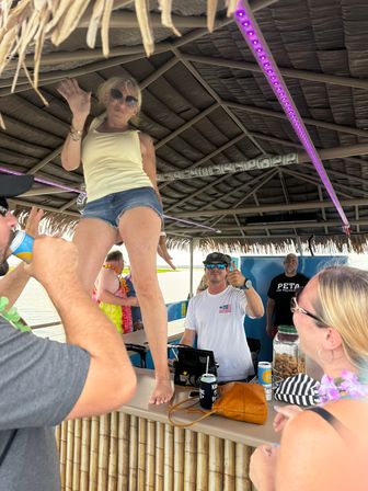 Barefoot woman dancing on a thatched-roof boat tiki bar counter while smiling patrons cheer and sip drinks at a waterfront party.