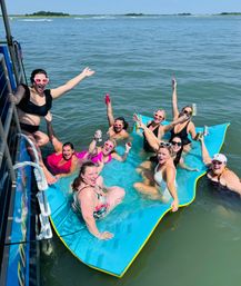 Group of smiling women in swimsuits lounging on a bright blue floating mat next to a boat, raising drinks and cheering in sunny coastal bay waters on a summer day.
