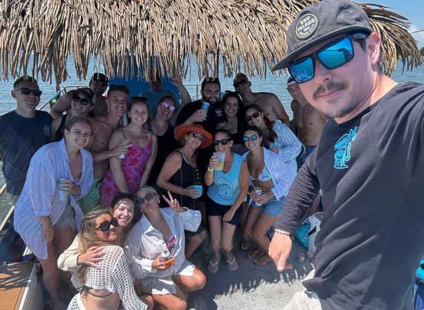 Large group of friends enjoying a sunny boat party under a thatched tiki canopy on calm coastal waters, wearing swimsuits and sunglasses and holding drinks.