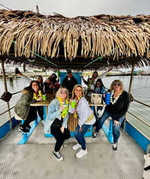 Group of friends on a thatched-roof tiki pontoon boat at a marina, wearing leis and novelty “50” glasses and holding colorful cups during a waterfront milestone birthday celebration.