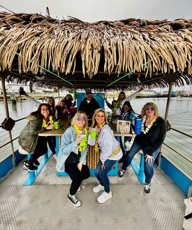 Group of friends on a thatched-roof tiki pontoon boat at a marina, wearing leis and novelty “50” glasses and holding colorful cups during a waterfront milestone birthday celebration.