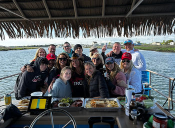 Group of friends and family posing on a tiki-roofed pontoon boat over a coastal marsh, smiling around trays of charcuterie, fruit and drinks with shoreline houses and a marina in the background.
