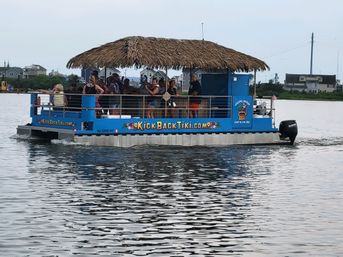 Blue tiki-style pontoon boat with a thatched roof carrying people on a sunny coastal bay cruise past waterfront homes