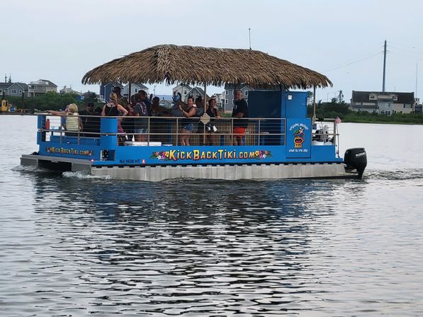 Blue tiki-style pontoon boat with a thatched roof carrying people on a sunny coastal bay cruise past waterfront homes