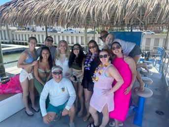 Group of friends smiling under a thatched tiki canopy on a pontoon boat at a sunny marina, wearing swimsuits, cover-ups, and leis.