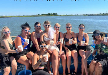 Group of women in swimsuits and heart-shaped sunglasses holding drinks on a sunny boat ride in a coastal bay — bachelorette party vibe with marshland and blue sky in the background.