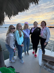 Five friends wearing floral leis and casual outfits posing on a boat deck under a thatched canopy during a colorful coastal bay sunset, calm water and cloud-filled sky in the background.
