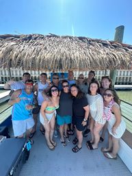 Smiling group of people in swimwear gathered under a thatched tiki roof on a sunny party boat at a coastal marina, dock and calm water visible