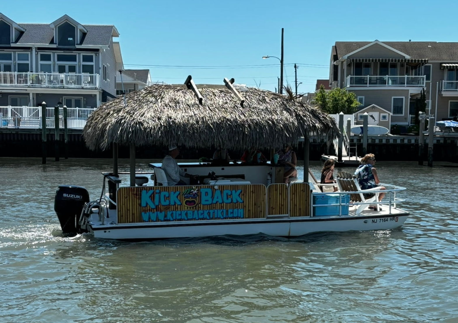 Thatched-roof tiki-style pontoon boat with outboard motor and passengers cruising a residential waterfront canal past multi-story coastal homes on a sunny summer day