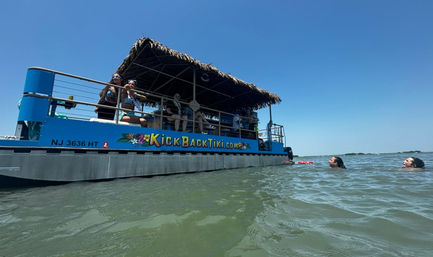 Bright blue tiki-style pontoon boat with a thatched roof anchored in shallow coastal water under a clear sky, passengers lounging on deck and two swimmers bobbing nearby.