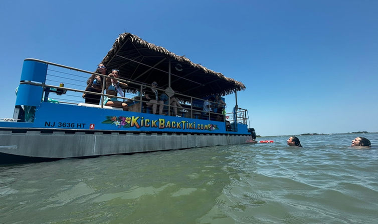 Bright blue tiki-style pontoon boat with a thatched roof anchored in shallow coastal water under a clear sky, passengers lounging on deck and two swimmers bobbing nearby.