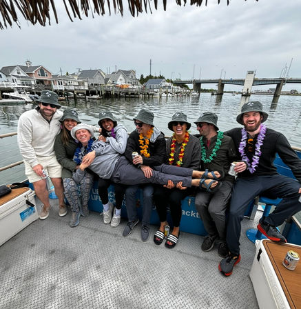 Group of friends wearing bucket hats and colorful leis laughing on a party boat at a coastal marina, one person held horizontally across laps with waterfront houses and a drawbridge in the background.