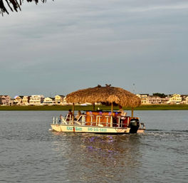 Tiki-style pontoon boat with a palm-thatched roof cruising a calm coastal inlet at golden hour, passengers aboard and pastel waterfront houses lining the shore.