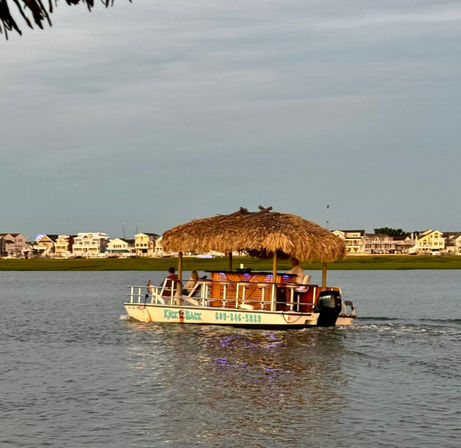 Tiki-style pontoon boat with a palm-thatched roof cruising a calm coastal inlet at golden hour, passengers aboard and pastel waterfront houses lining the shore.