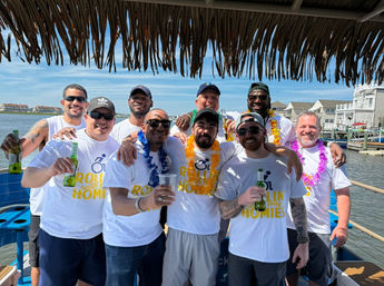 Group of men in matching Rollin with my homies T-shirts wearing leis and sunglasses, holding beers on a sunny boat at a coastal marina under a thatched tiki canopy