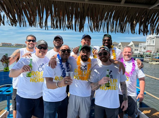 Group of men in matching Rollin with my homies T-shirts wearing leis and sunglasses, holding beers on a sunny boat at a coastal marina under a thatched tiki canopy