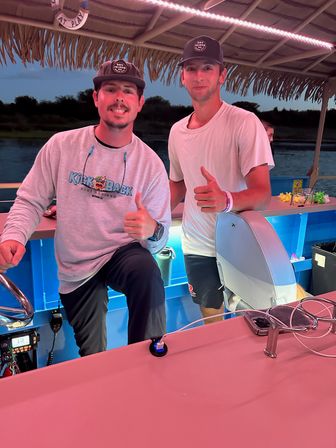 Two men giving thumbs-up on a tiki-style pontoon boat at dusk with pink LED lighting, water and a tree-lined shore in the background.